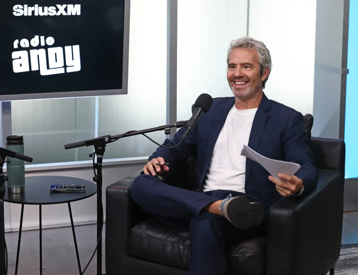 Andy Cohen sits in an armchair with his leg crossed. There is a microphone in front of him, and he holds a paper in his hand.