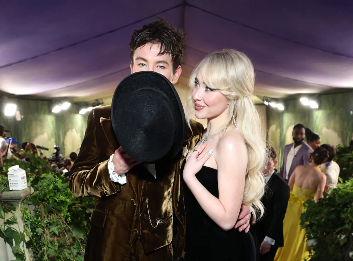 Barry Keoghan covers his face with a hat. Sabrina Carpenter wears a black dress and stands with him.