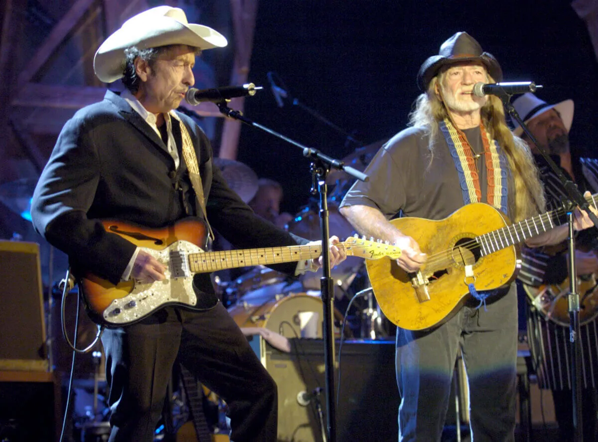 Bob Dylan and Willie Nelson play guitars and stand in front of microphones.