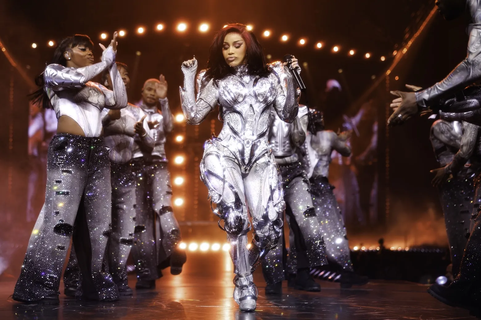 Cardi B in a full-body suit dancing on the 'Little Miss Drama Tour' with backup dancers around her.