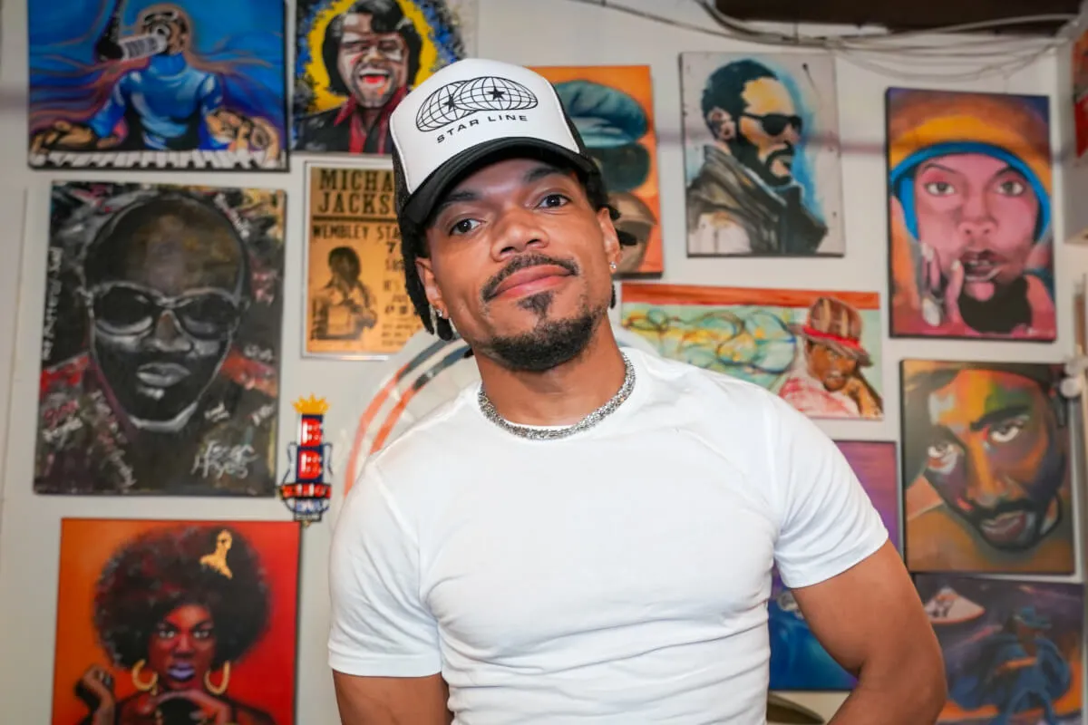 Chance the Rapper wears a white shirt and a hat. He stands in front of a wall covered in pictures.