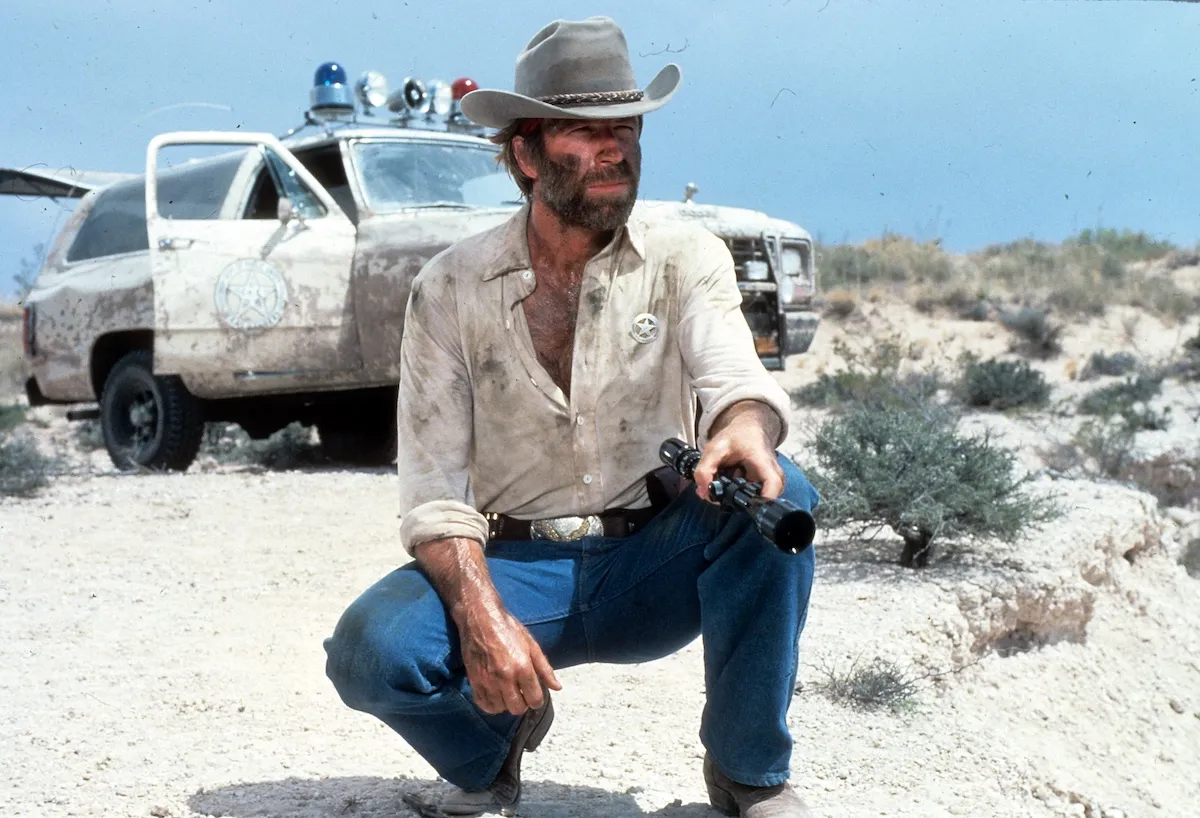 Chuck Norris, wearing a cowboy hat and holding a flashlight while crouching in the desert, in 'Lone Wolf McQuade