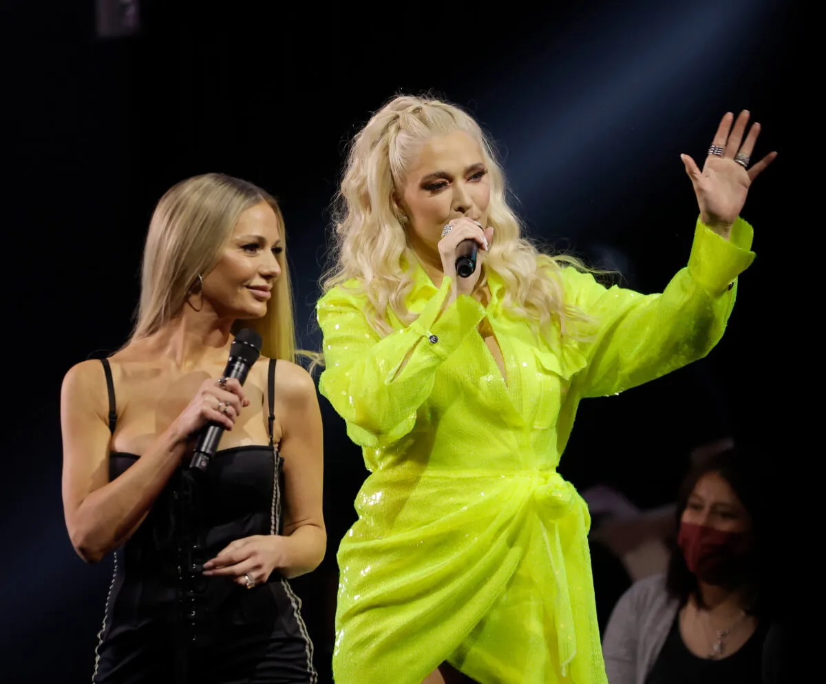 Dorit Kemsley wears a black dress and stands with Erika Jayne, who wears yellow. They both hold microphones.