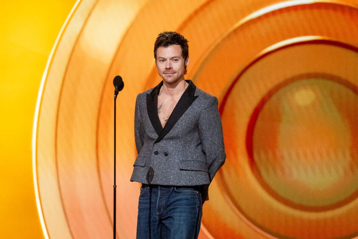 Harry Styles wears a gray cropped blazer and stands in front of a microphone.