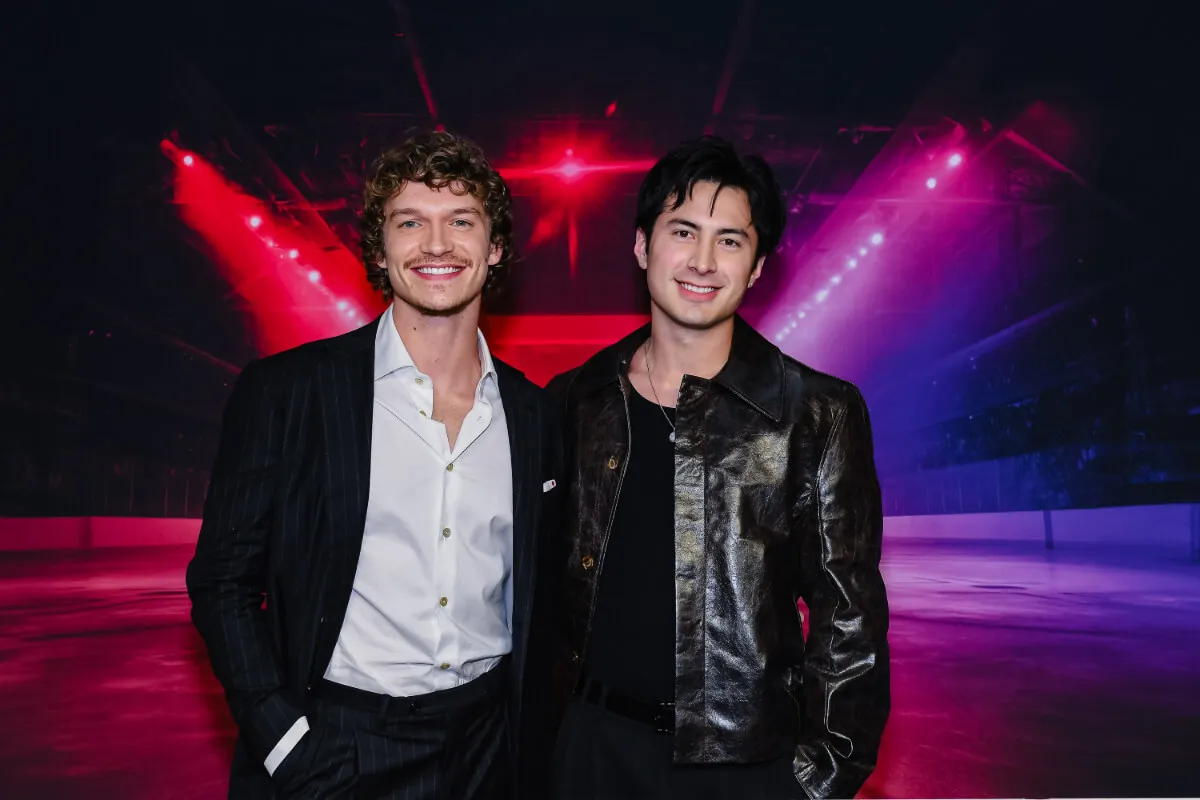 'Heated Rivalry' stars Connor Storrie and Hudson Williams stand together. Storrie wears a white shirt and black jacket, and Williams wears a black shirt and black jacket.