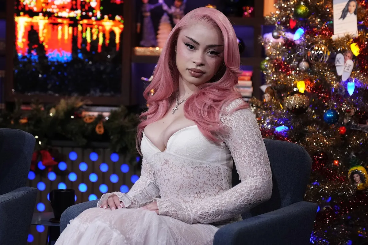 Ice Spice looking toward the camera on 'Watch What Happens Live.' She's sitting on a couch in a long-sleeve light pink dress, and her pink hair is cascading down her shoulders.