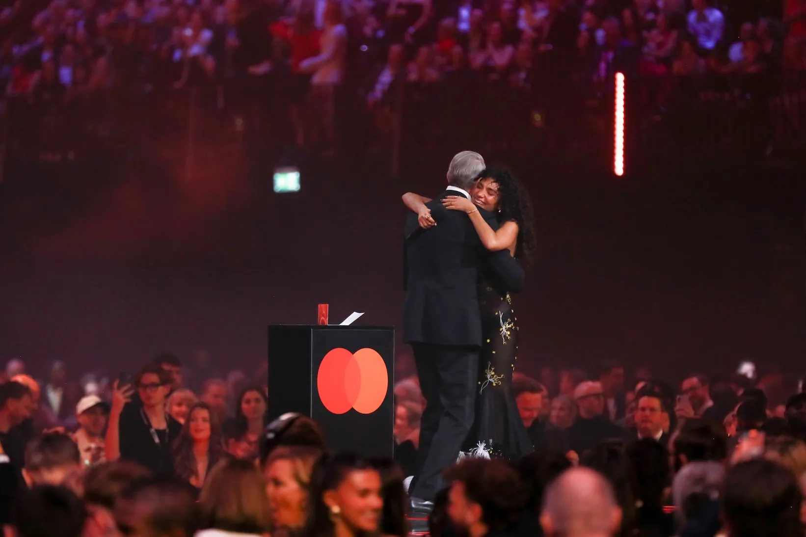 Jeff Goldblum and Olivia Dean embracing onstage at the BRIT Awards in 2026. A crowd is below them.