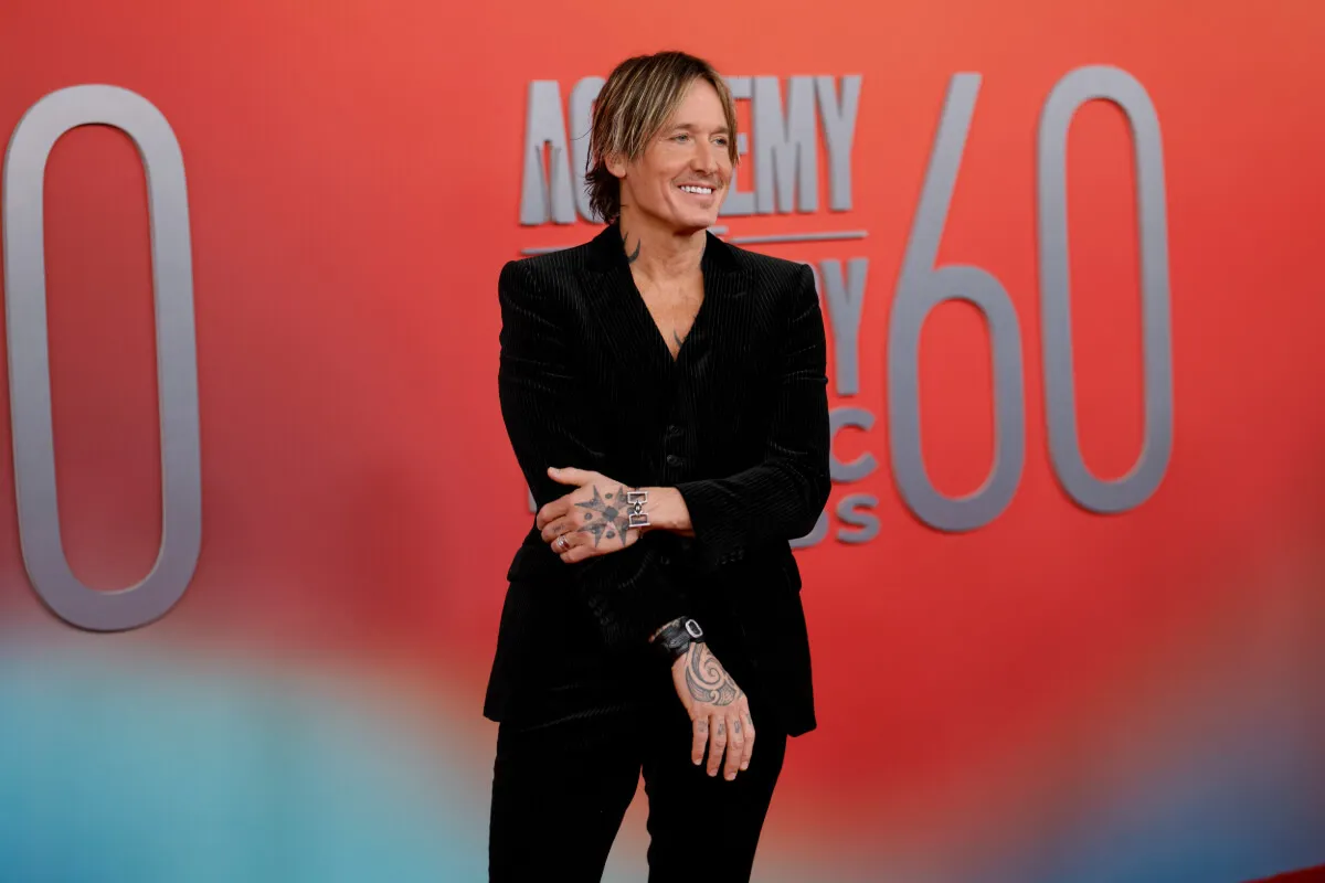 Keith Urban wears a black suit and stands in front of an orange background.