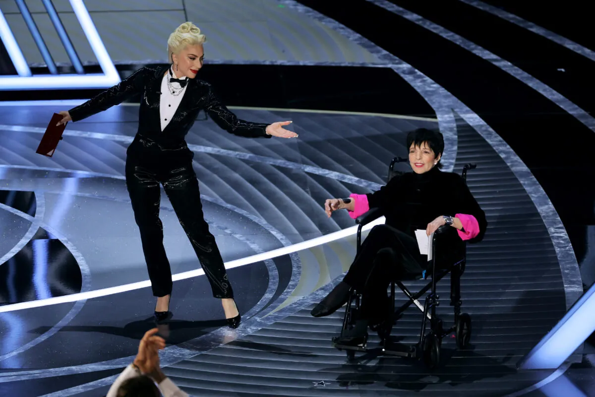 Lady Gaga wears a tuxedo and gestures to Liza Minnelli, who wears black with pink sleeves and sits in a wheelchair. They are presenting at the Oscars.