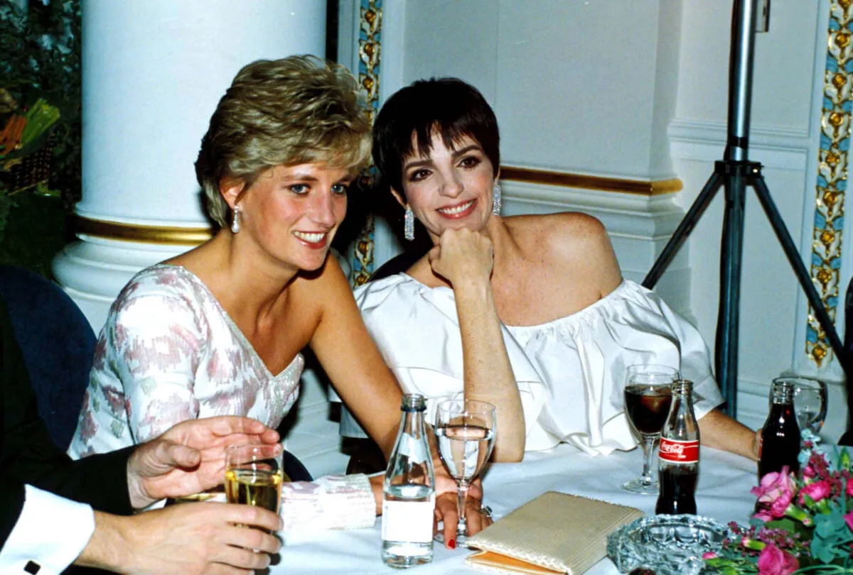 Liza Minnelli and Princess Diana wear white and sits at a table together. They smile.