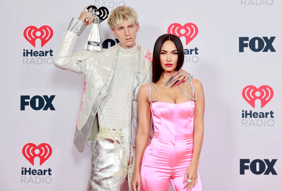 MGK wears white and stands with Megan Fox, who wears a pink jumpsuit.