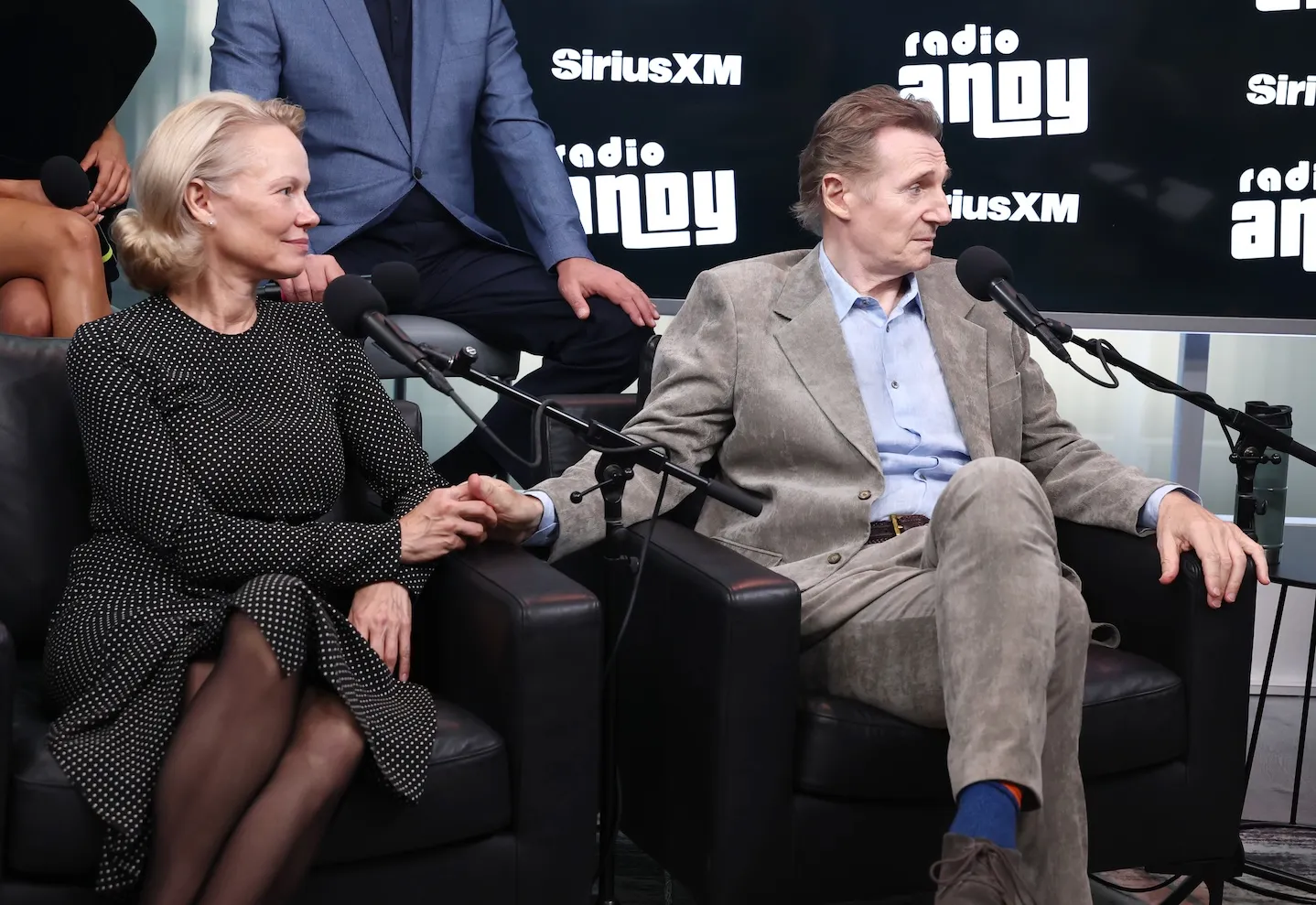 Pamela Anderson and Liam Neeson holding hands while sitting in chairs at SiriusXM Studios for an interview
