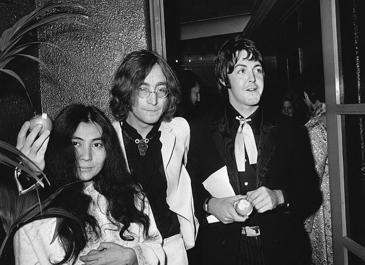 A black and white picture of Yoko Ono, John Lennon, and Paul McCartney. Lennon and McCartney hold apples.
