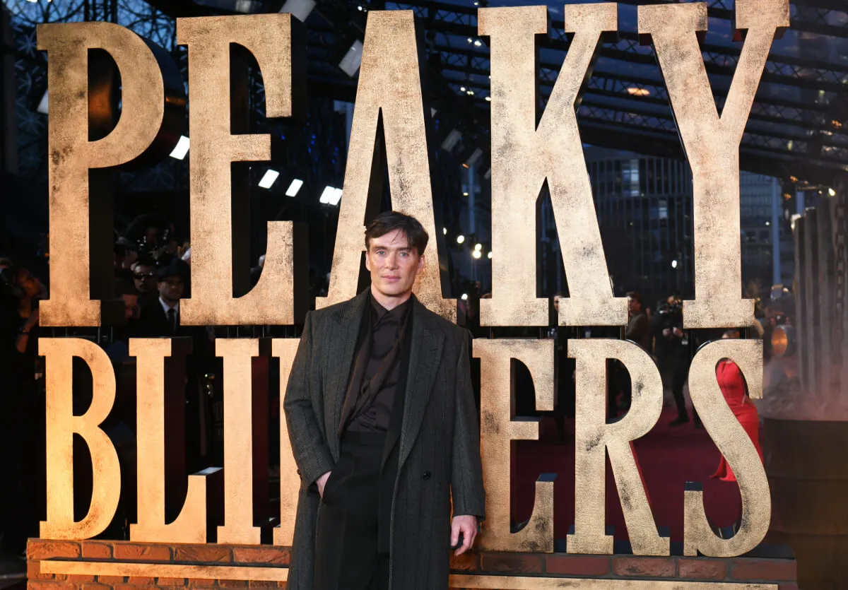 Cillian Murphy wears a black suit and stands in front of a 'Peaky Blinders' sign.