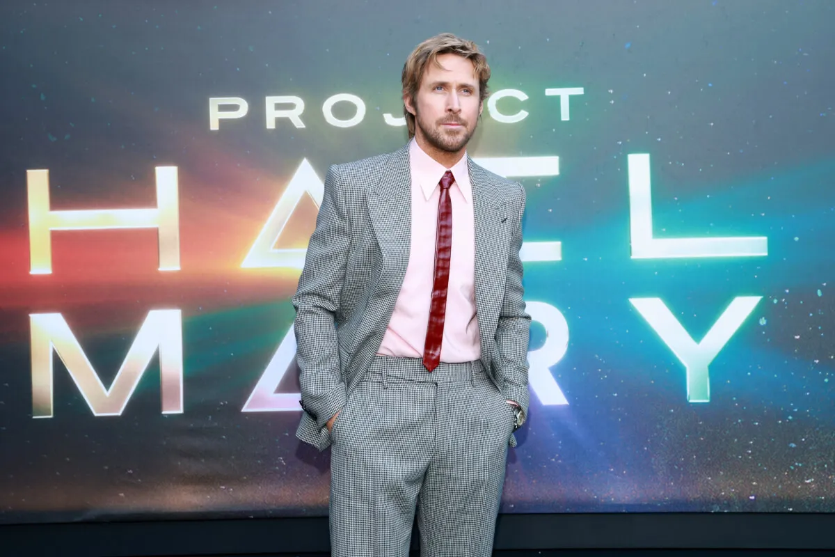 Ryan Gosling wears a checkered suit and red tie. He stands in front of a sign for 'Project Hail Mary.'