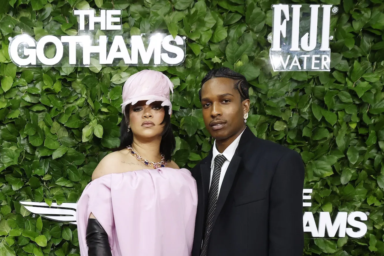 Rihanna in an all-pink outfit and a pink bucket hat posing next to A$AP Rocky in a suit against a green leafy background for an event