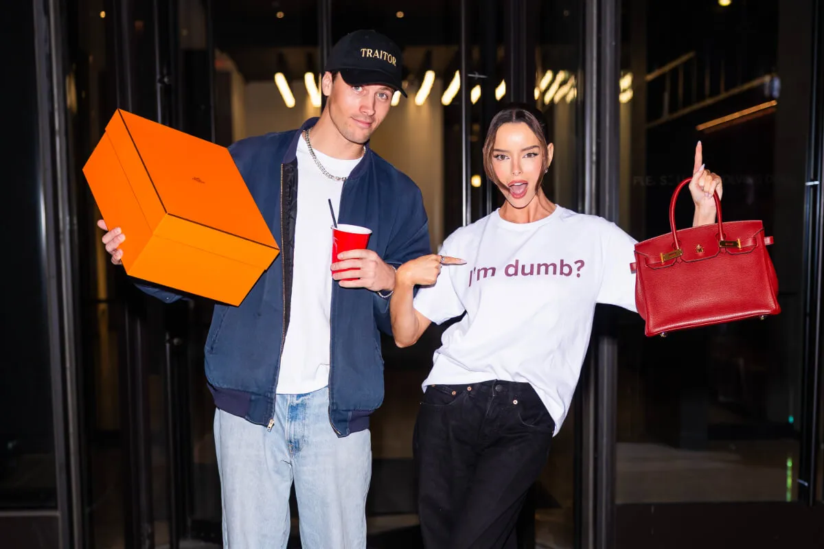 Rob Rausch holds an orange box in one hand and a red solo cup in the other. Maura Higgins points to her shirt, which reads "I'm Dumb'?" She carries a red Birkin bag in the other hand.