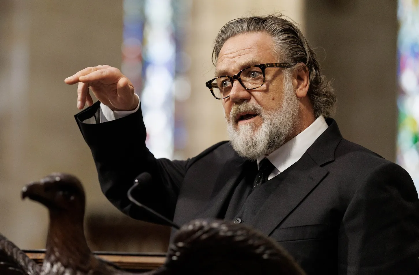 Russell Crowe reading a eulogy with his right hand up mid-sentence. He's wearing a black suit.