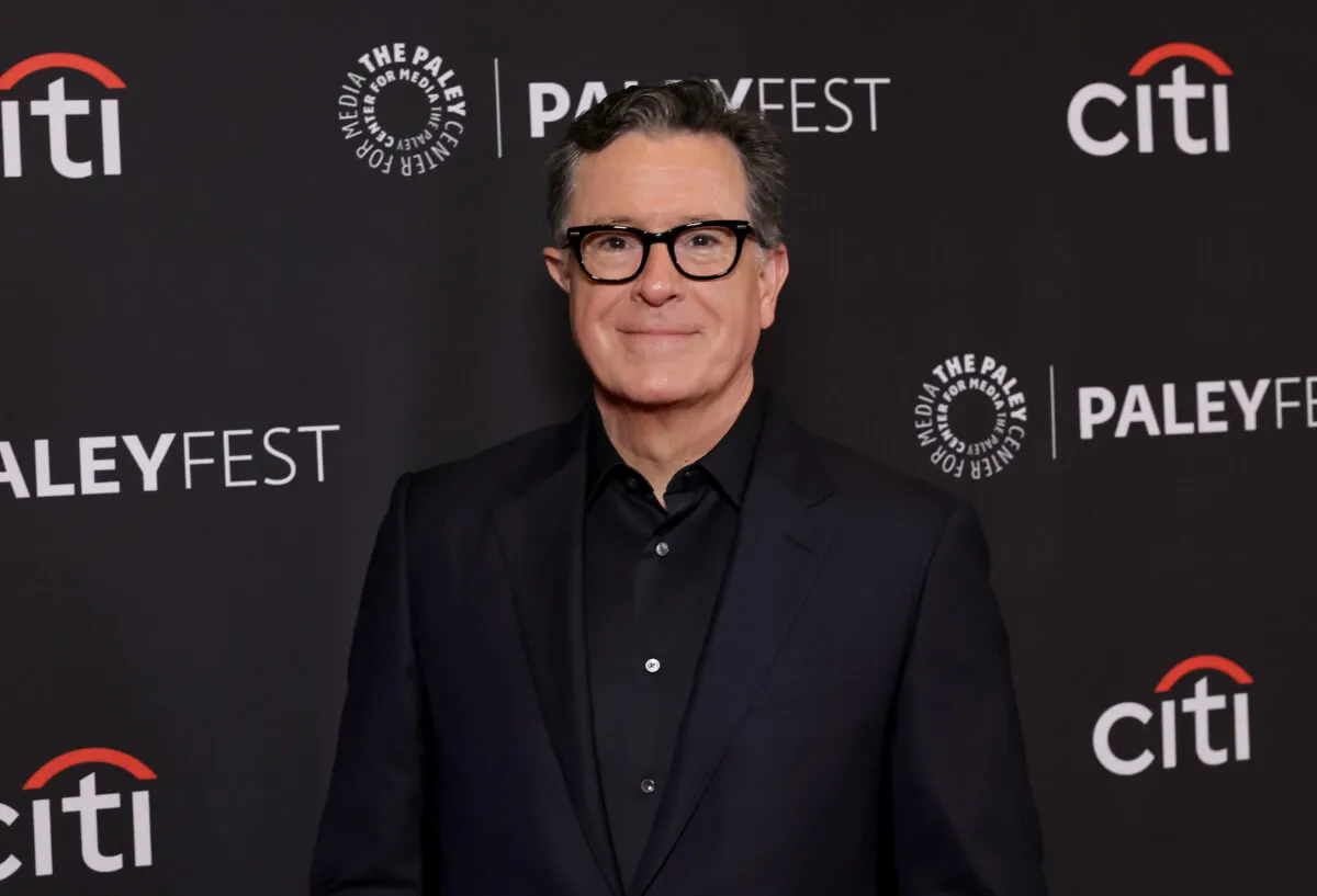 Stephen Colbert wears glasses and a black shirt and jacket.