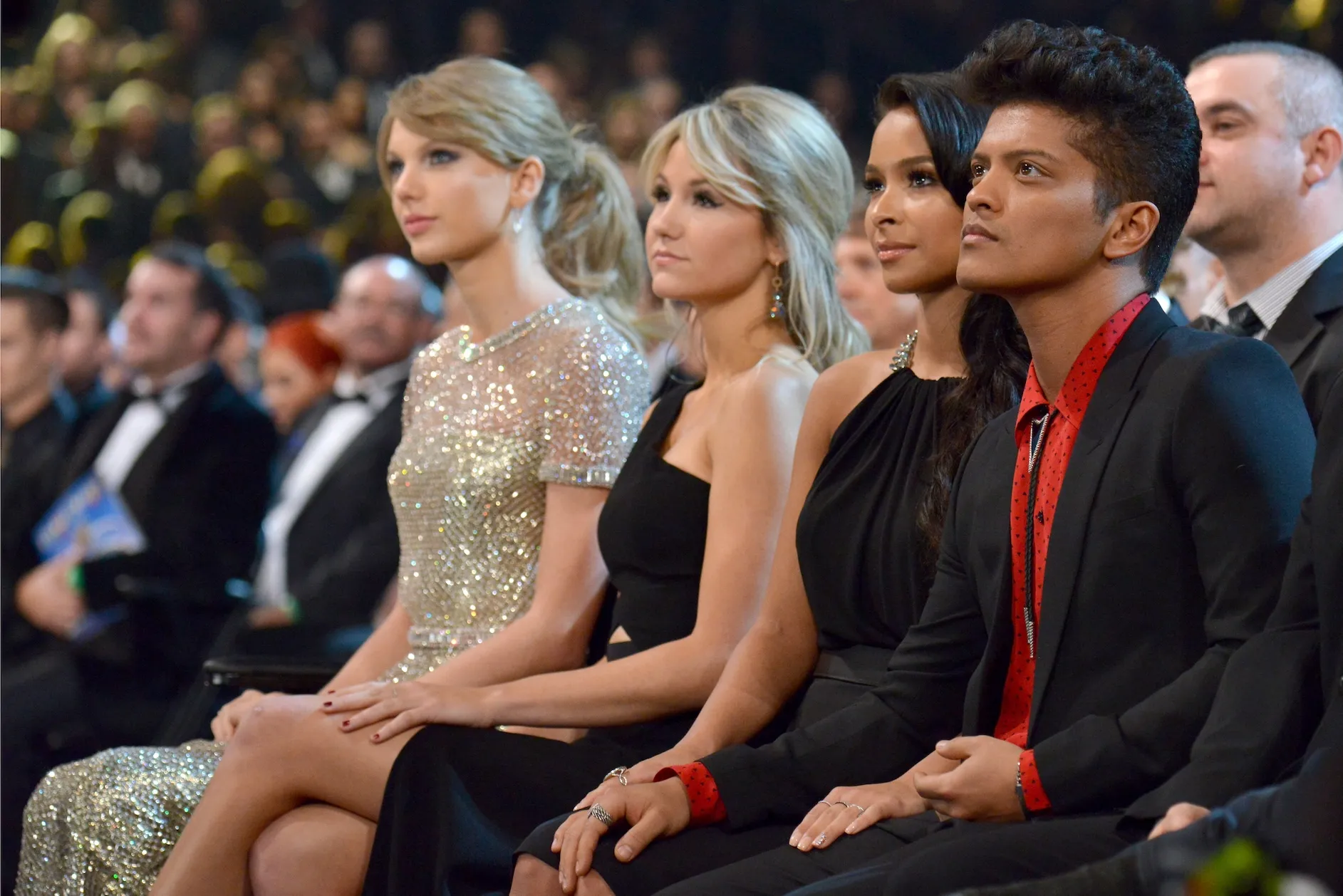Taylor Swift and Bruno Mars attend the 56th GRAMMY Awards. They are sitting in a row near each other and focused on the presenters.