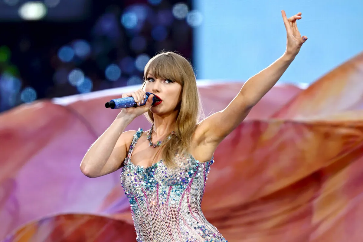 Taylor Swift wears a sparkly bodysuit and sings into a microphone. She holds up one arm.