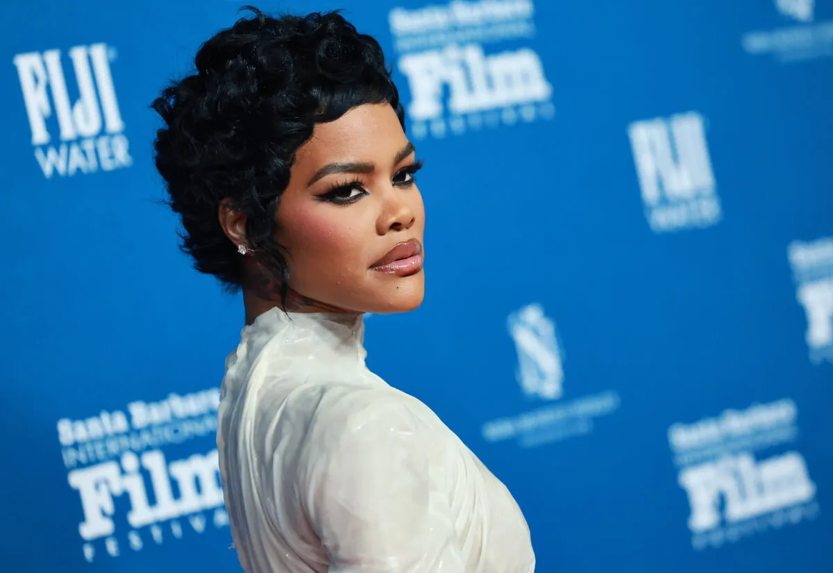 Teyana Taylor wears a white dress and stands in front of a blue background.