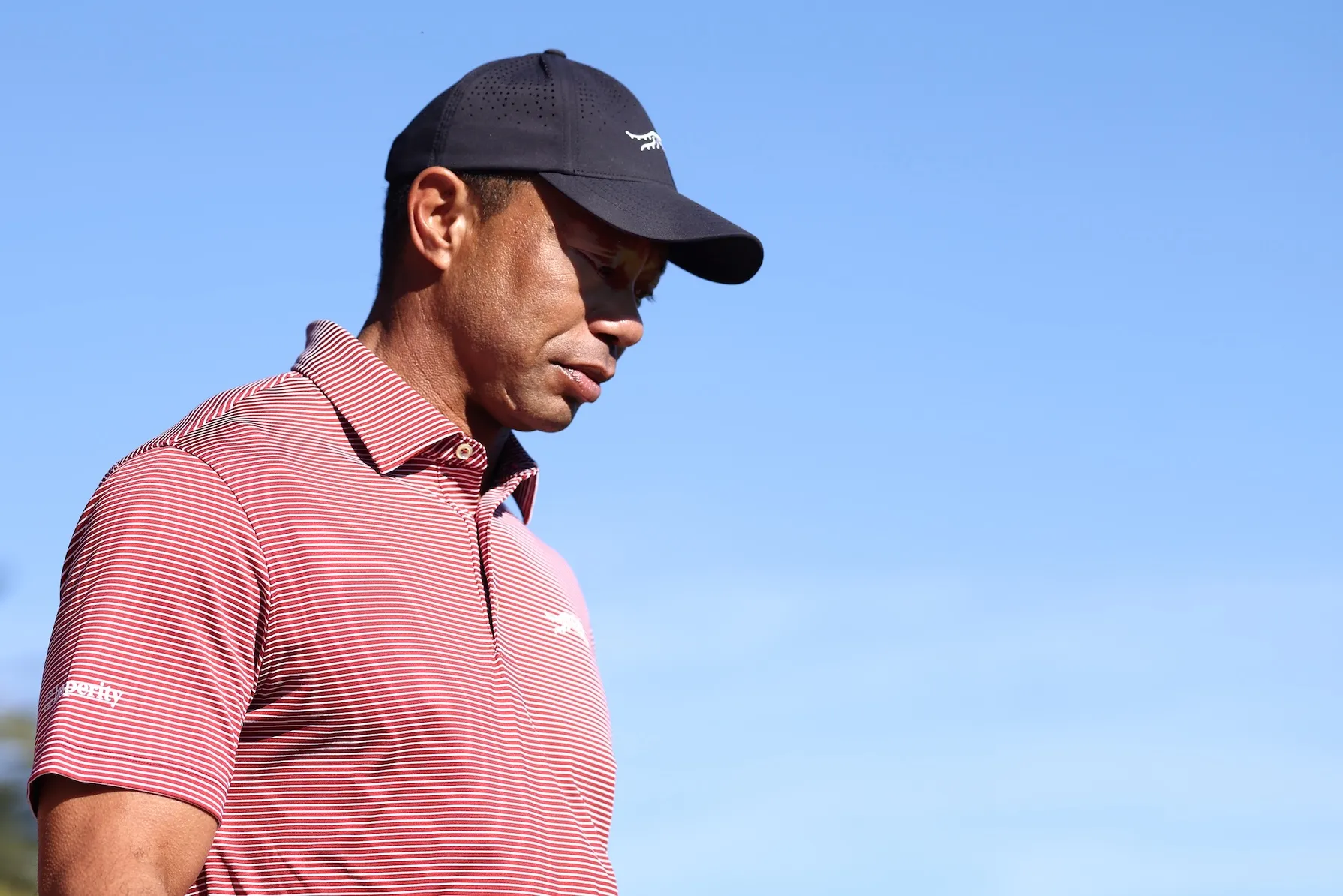 Tiger Woods in a red striped polo shirt and a baseball cap looking forward while playing golf