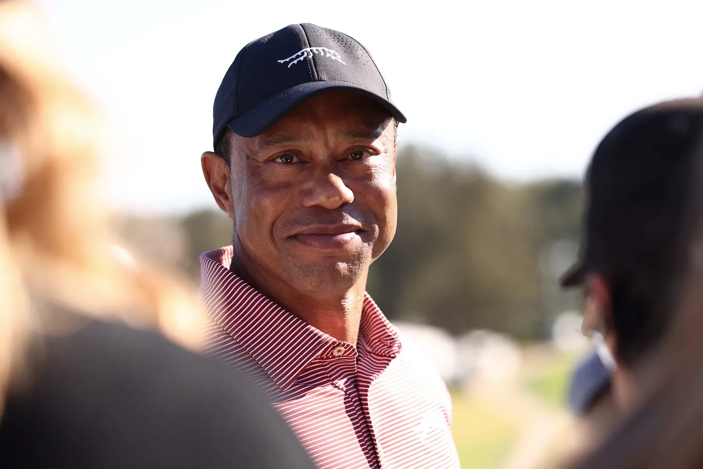 A close-up of Tiger Woods slightly smiling while wearing a hat and a button-up shirt during a golf tournament.