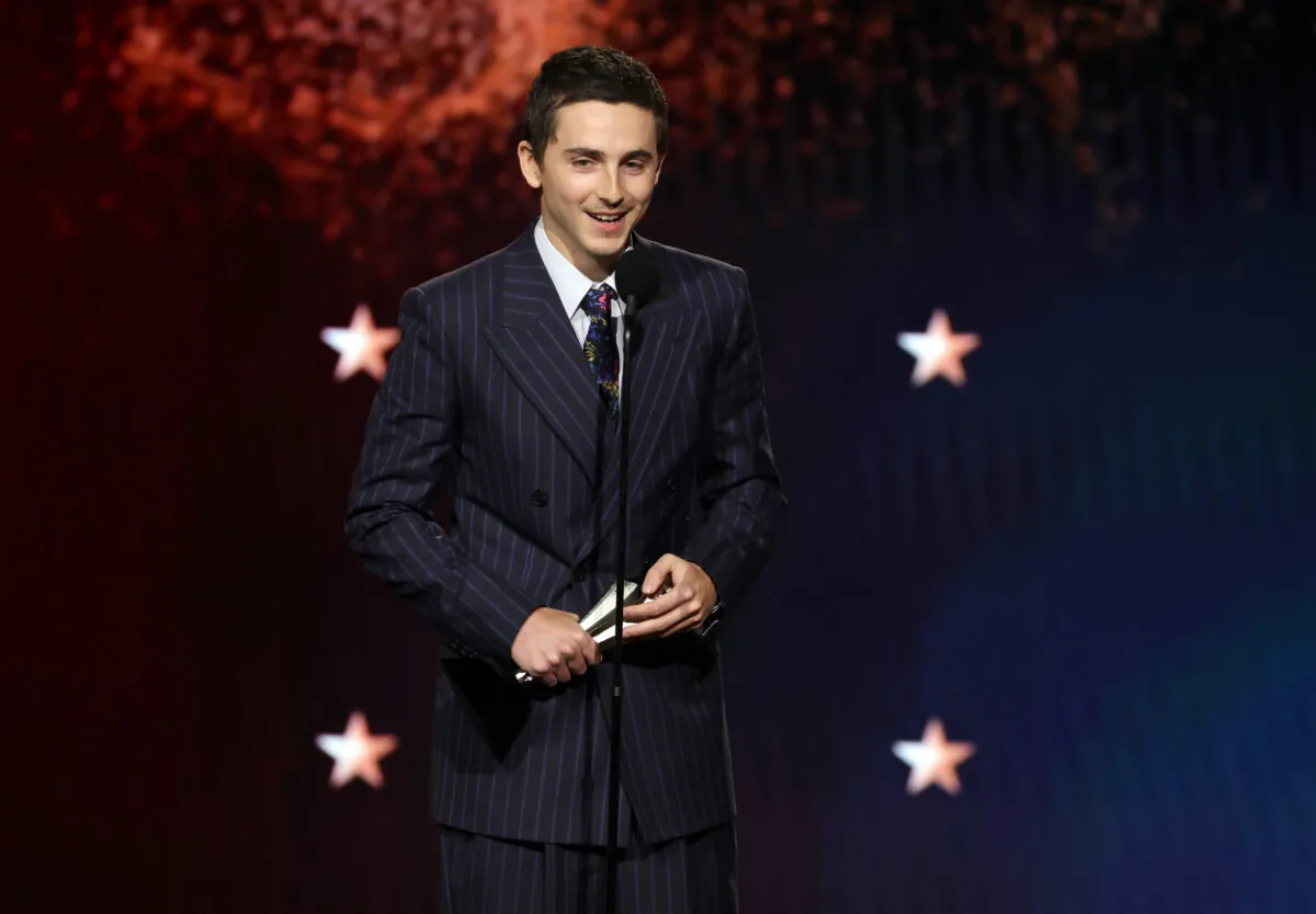 Timothee Chalamet wears a suit and stands in front of a microphone.