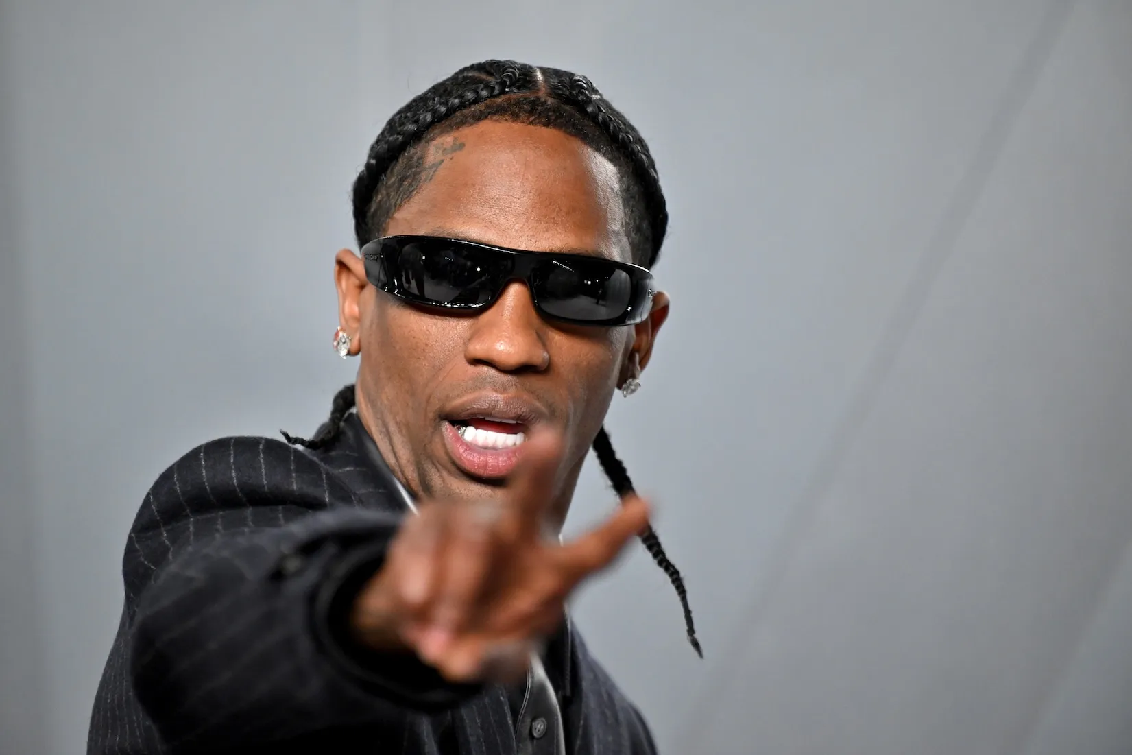 A close-up of Travis Scott flashing a peace sign at the Vanity Fair Oscar Party in 2026. He's wearing a suit and sunglasses.