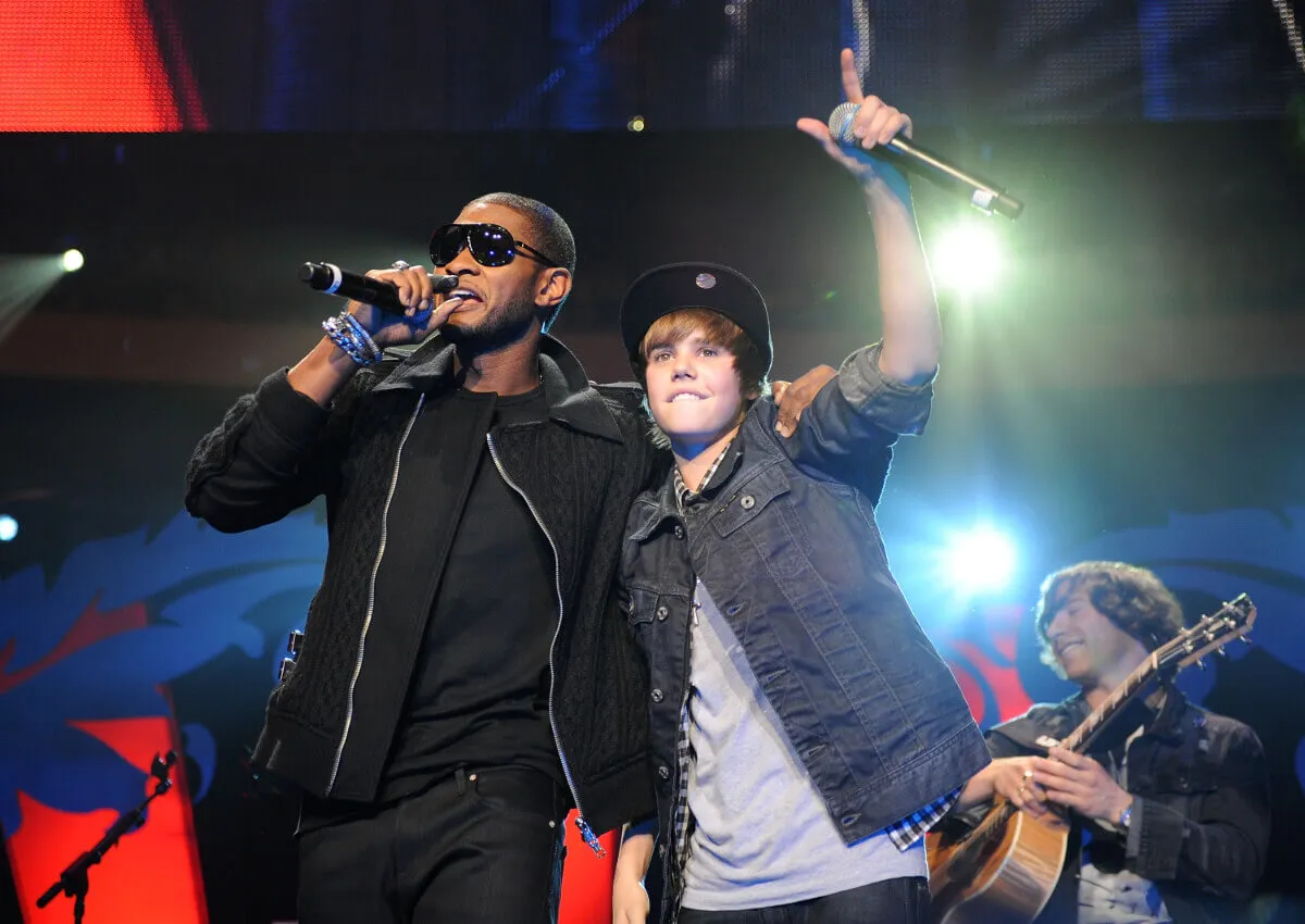 Usher and Justin Bieber stand with their arms around each other on a stage. Usher wears sunglasses and Bieber wears a hat. They both hold microphones.