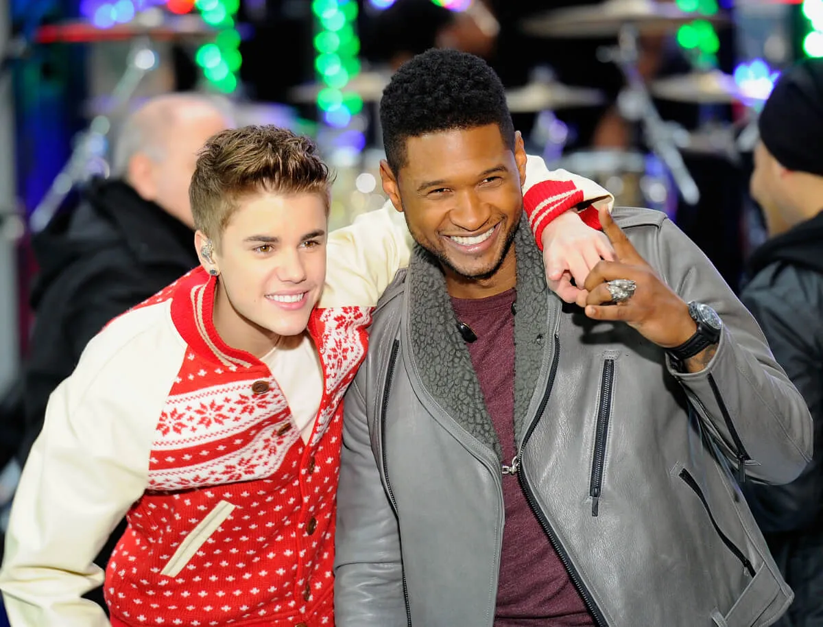Justin Bieber wears a red and white jacket. He stands with his arm around Usher's shoulders. Usher wears a grey jacket.