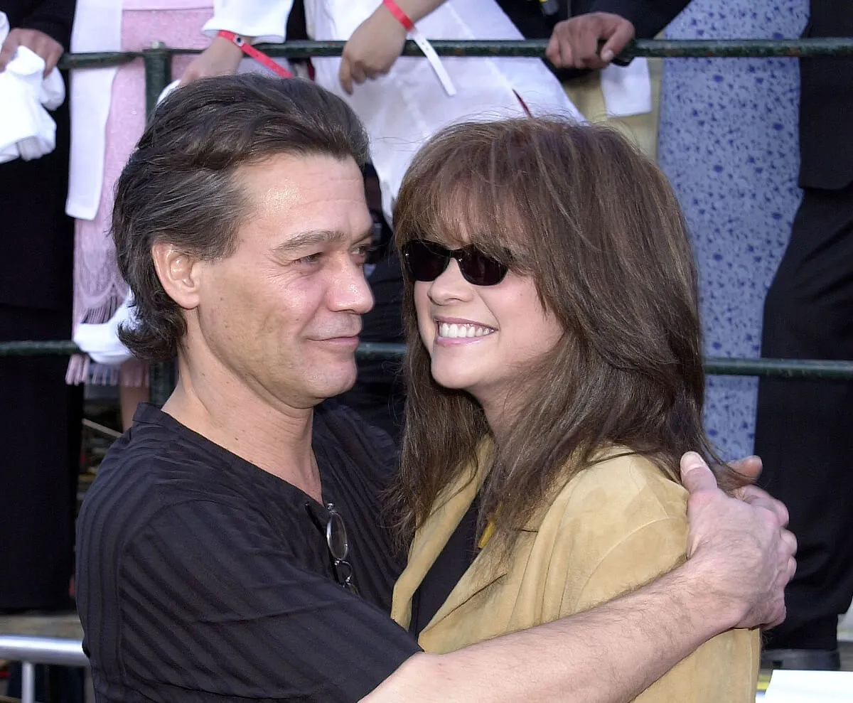 Eddie Van Halen wears a black shirt and embraces Valerie Bertinelli. She wears a yellow jacket and sunglasses.
