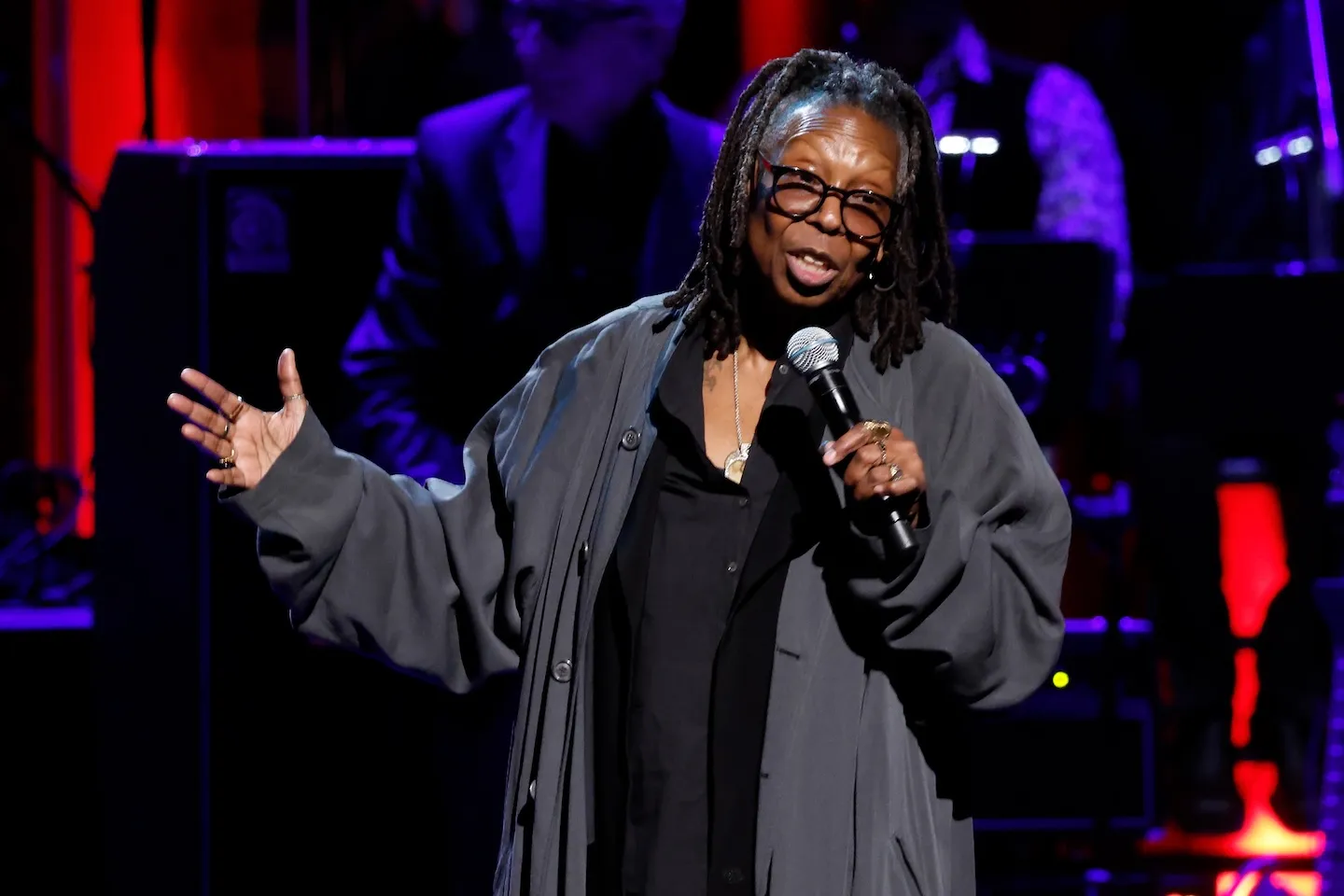 Whoopi Goldberg performing onstage with a microphone up to her mouth. She's gesturing with one hand.