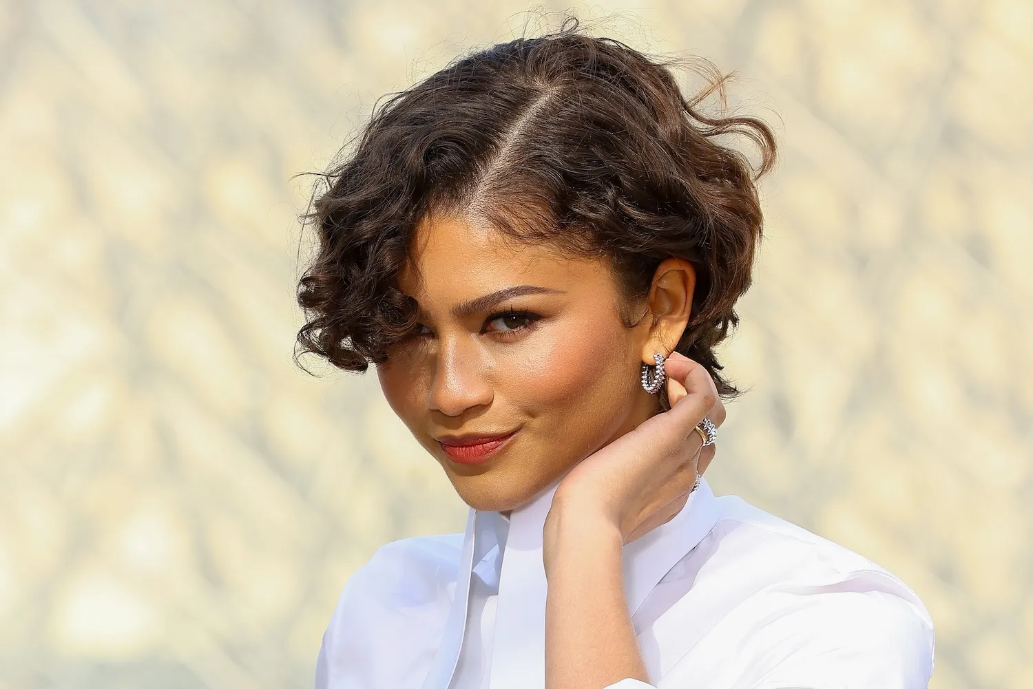 A close-up of Zendaya as she tucks hair behind her ear at a fashion show. She's wearing a structured white dress.