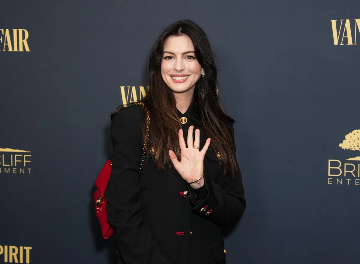 Anne Hathaway wears black with a red purse. She waves.