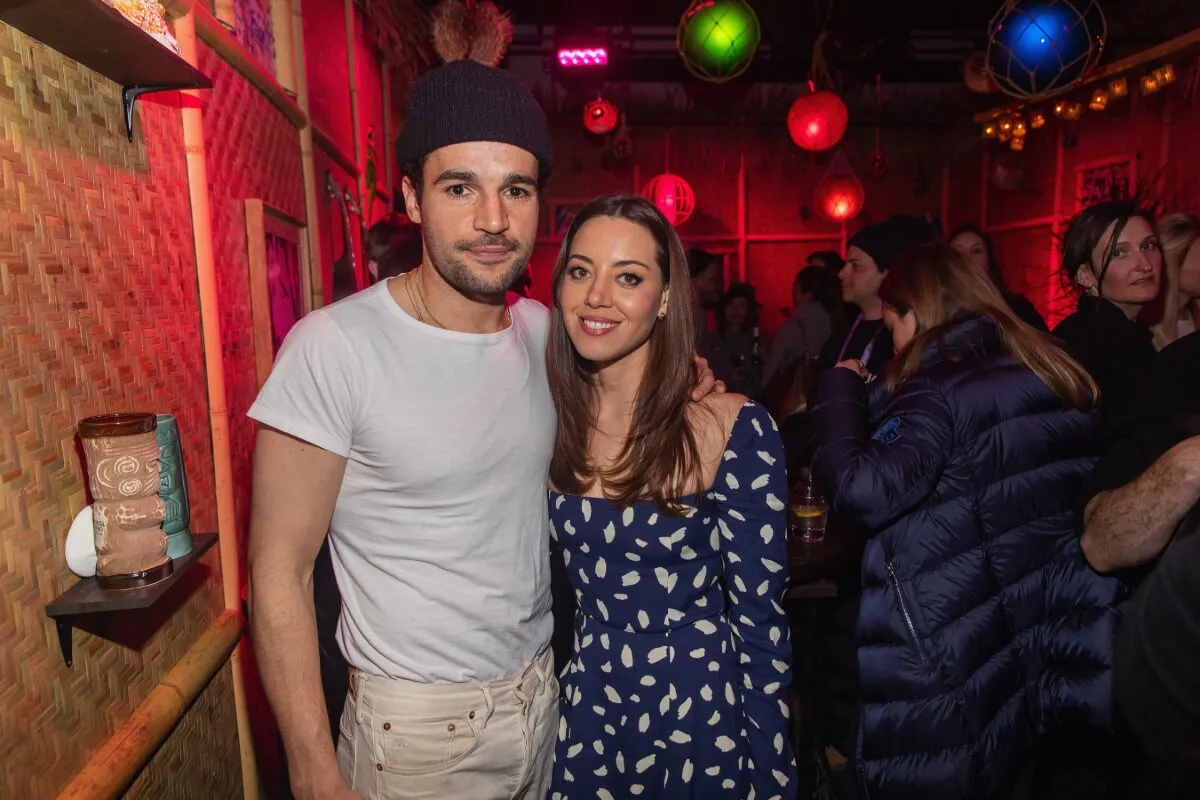 Christopher Abbott wears a white shirt and a hat and stands with Aubrey Plaza. She wears a blue and white dress.