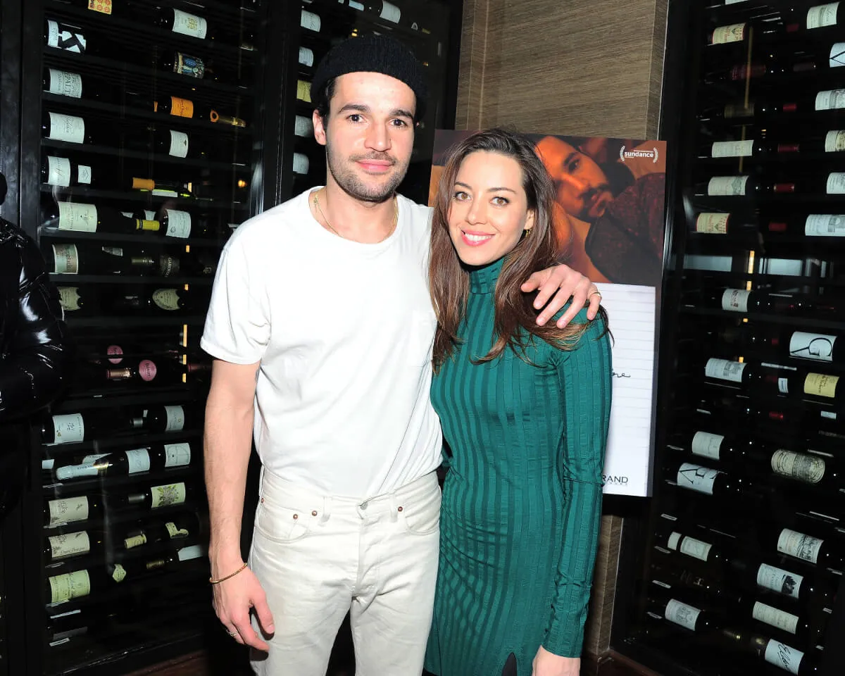 Christopher Abbott wears a white shirt and white pants with a black hat. He stands with his arm around Aubrey Plaza's shoulders. She wears a teal dress.