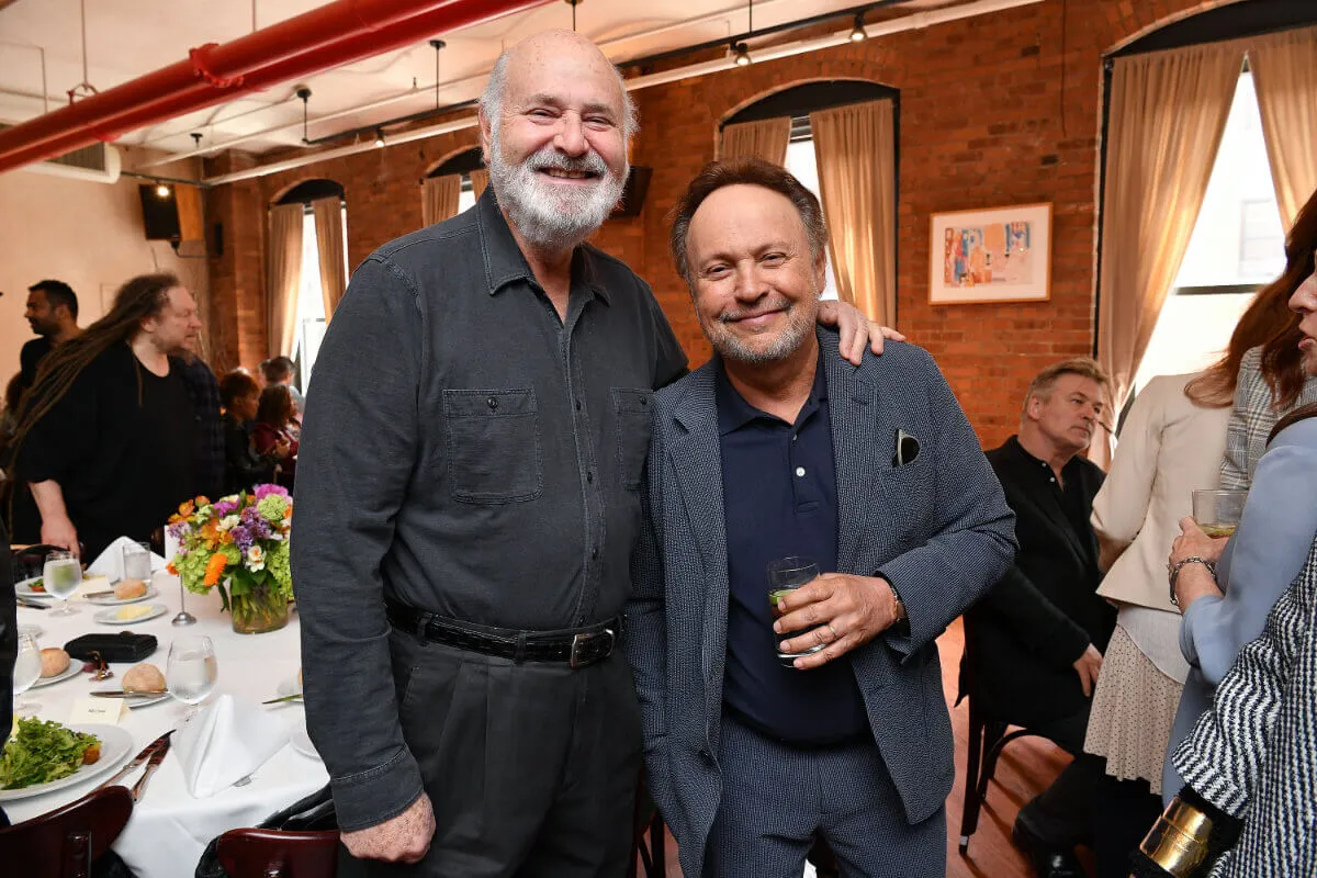 Rob Reiner wears a gray shirt and stands with his hand on Billy Crystal's shoulder. Crystal wears a blue suit.