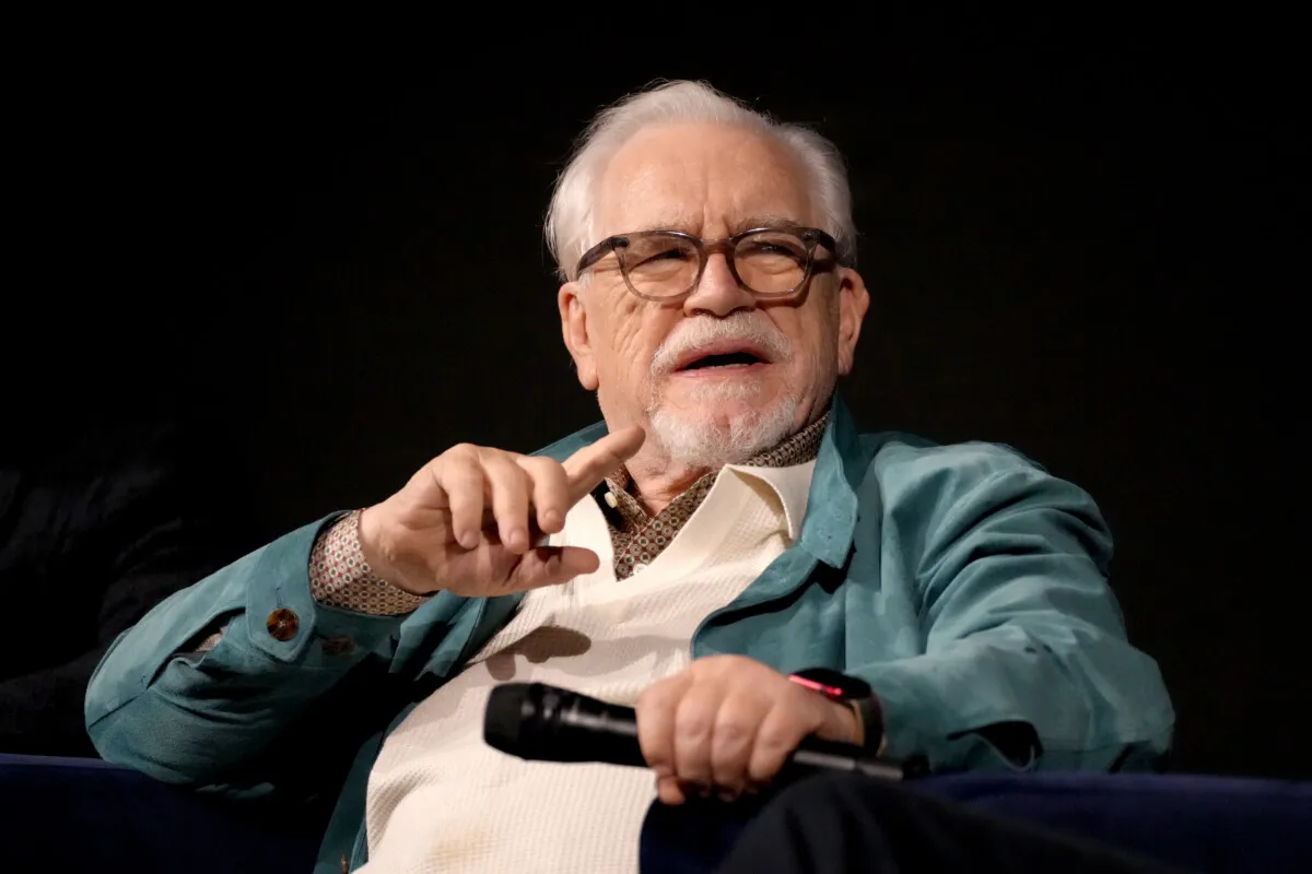 Brian Cox wears a blue jacket and glasses. He holds a microphone.