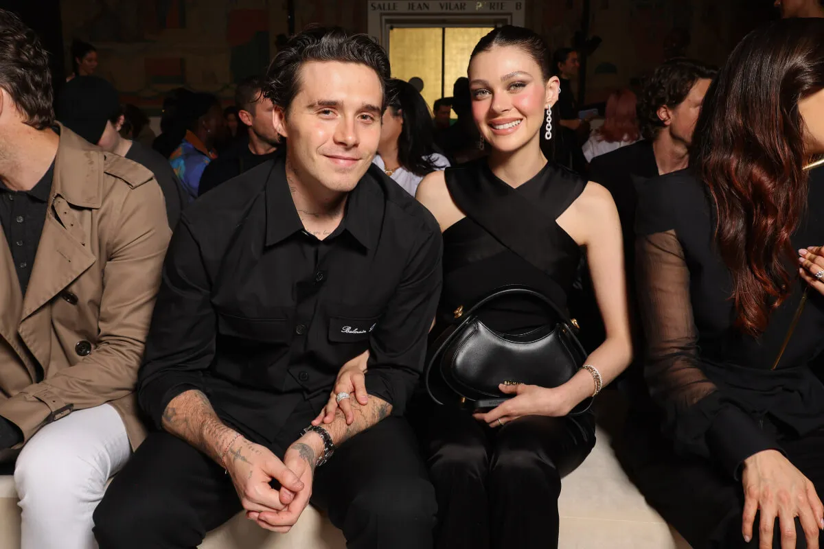 Nicola Peltz wears a black dress and sits with Brooklyn Beckham, who wears a black shirt.