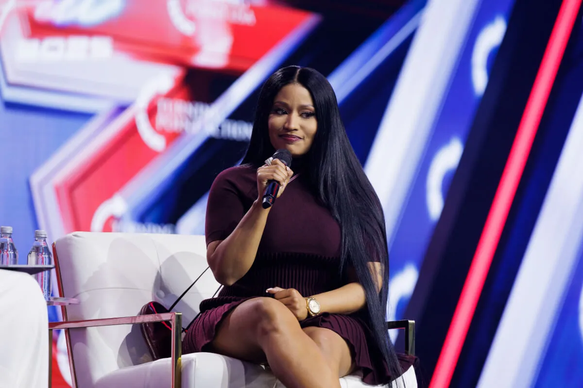 Nicki Minaj wears a purple dress and sits in an arm chair. She holds a microphone.
