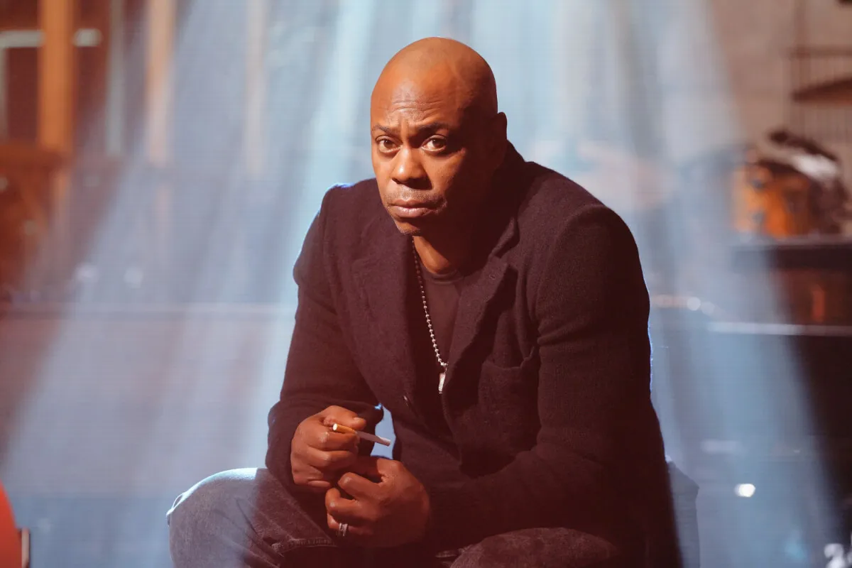 Dave Chappelle wears black and sits on a stage.