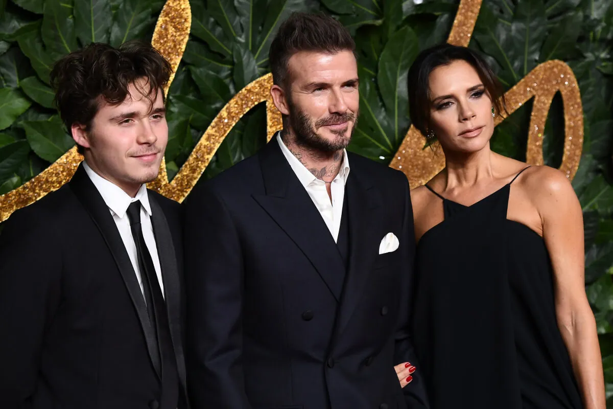 Brooklyn, David, and Victoria Beckham wear black. They stand against a green and gold background.