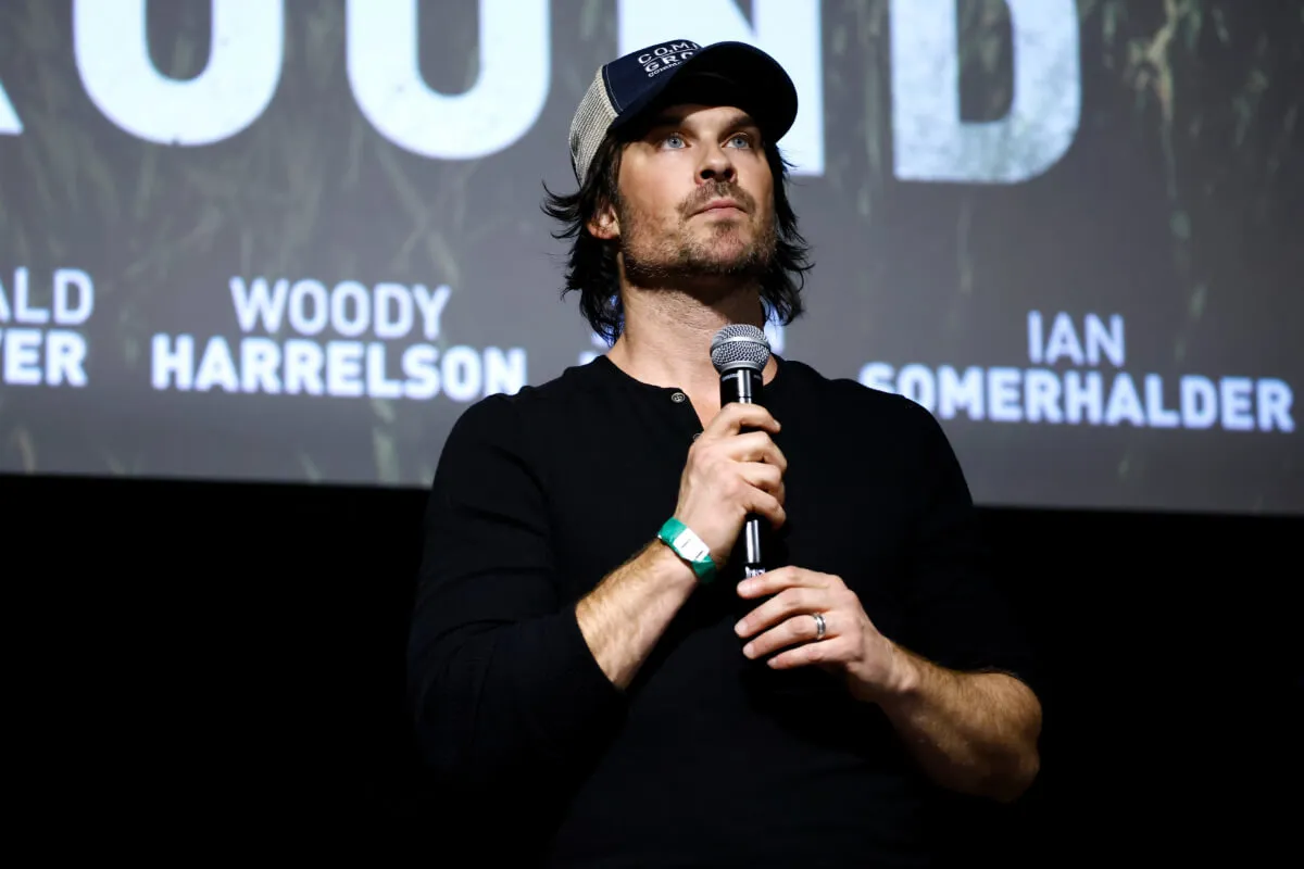 Ian Somerhalder wears a baseball hat and holds a microphone.