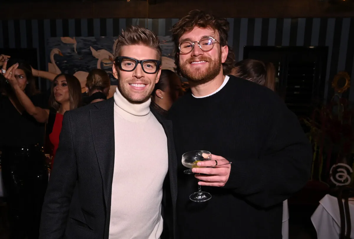 Kyle Cook wears a turtleneck and stands with West Wilson, who wears a black sweater. They both wear glasses, and Wilson holds a drink.