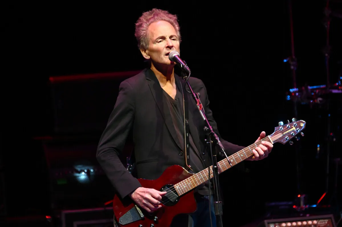 Lindsey Buckingham wears black and plays guitar in front of a microphone.