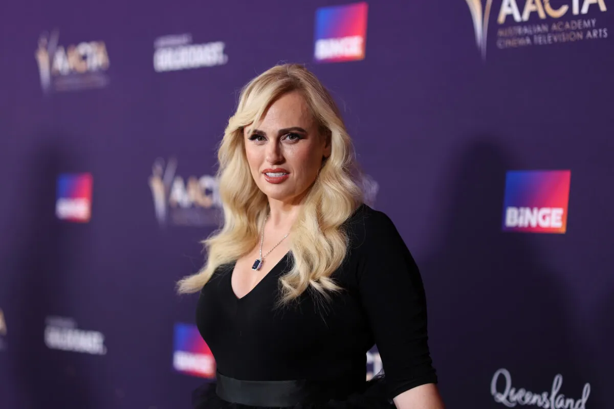 Rebel Wilson wears black and stands in front of a purple background.