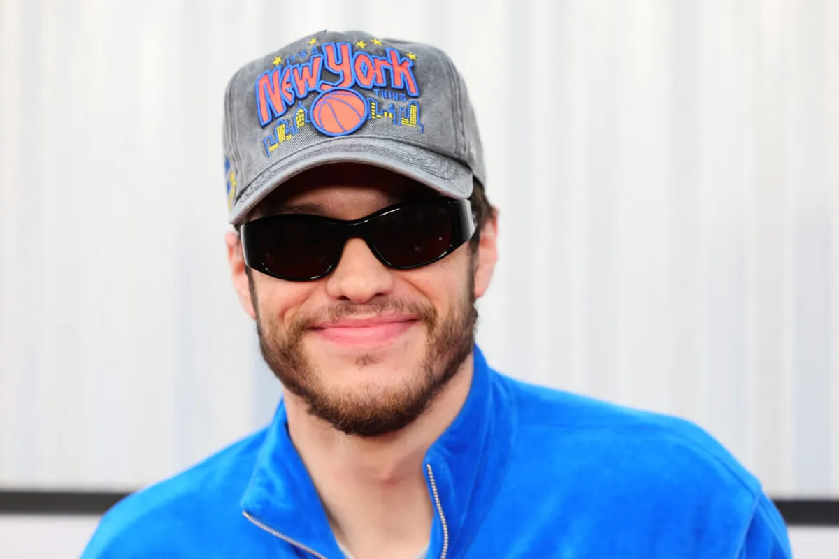 Pete Davidson wears a blue jacket, sunglasses, and a New York hat.