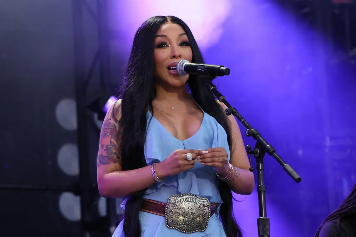 K. Michelle wears a blue dress with a large belt. She stands in front of a microphone.